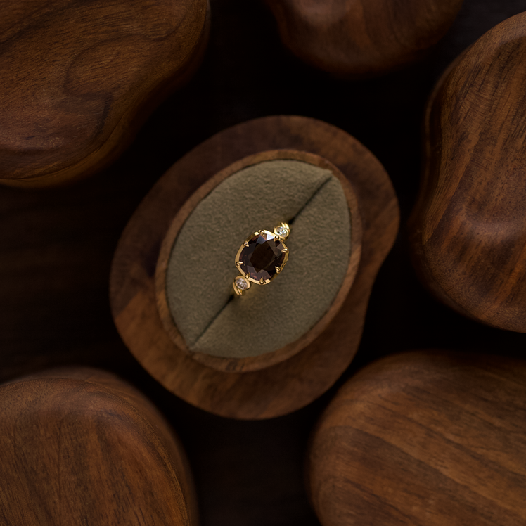 A gold ring with a dark burgundy gemstone displayed on a circular green velvet ring stand, set within a carved wooden tray and surrounded by other wooden pieces on a dark background.