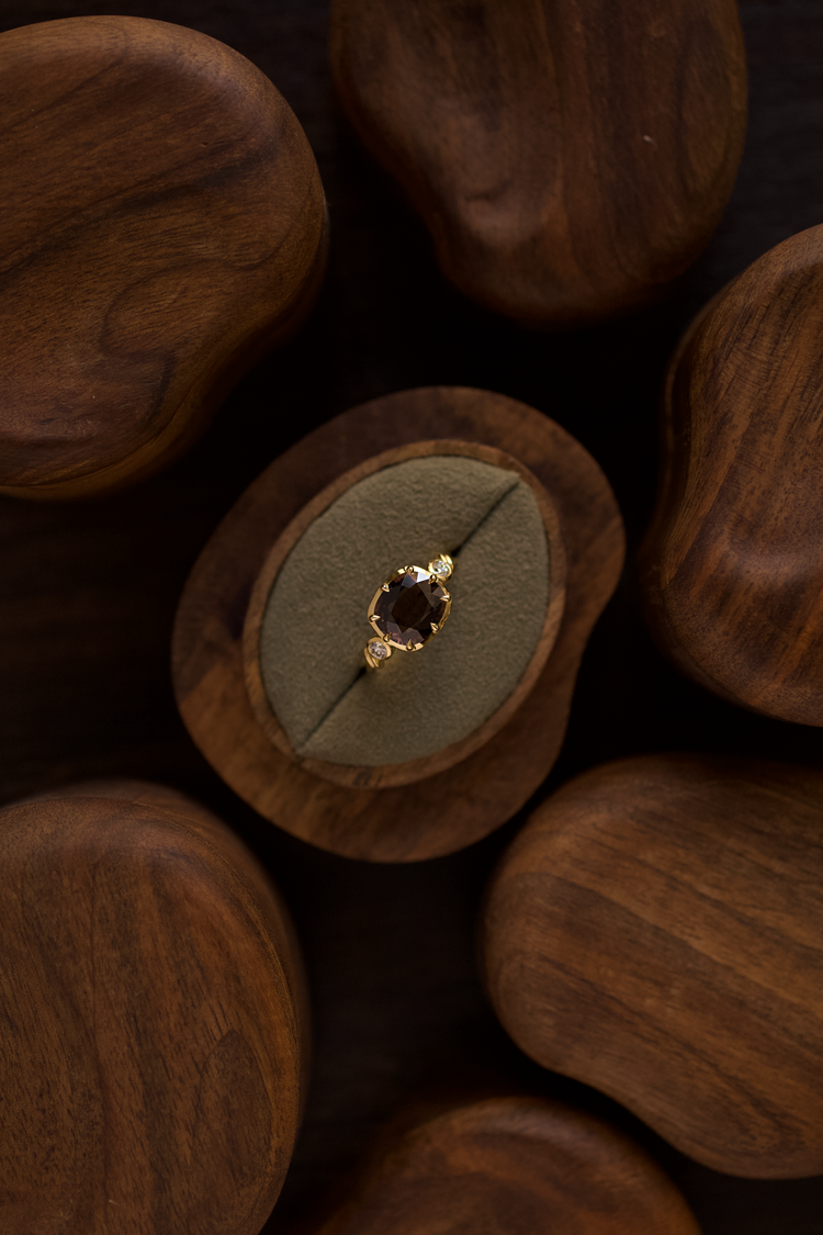 A gold ring with a dark burgundy gemstone displayed on a circular green velvet ring stand, set within a carved wooden tray and surrounded by other wooden pieces on a dark background.