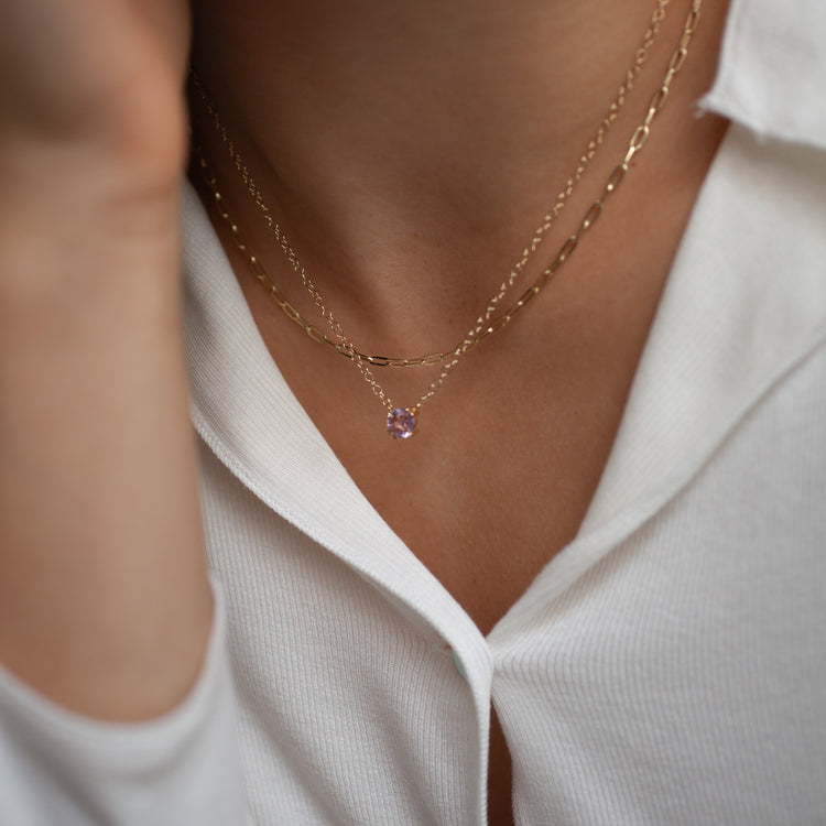 Close-up of a person wearing a gold necklace with a pink gemstone against a white shirt.